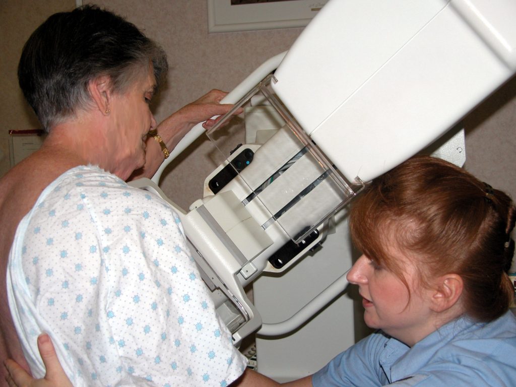 ”US_Navy_021025-N-6498N-001_Mammogram_technician,_aids_a_patient_in_completing_her_annual_mammogram_evaluation.jpg” by U.S. Navy photo by Ensign Ann-Marie Al Noad and is in the Public Domain. Photo showing a technician adjusting a patient for their mammogram
