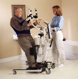 "invacare-reliant-350-electric-sit-to-stand-lift-3.jpg" by Invacare. Used under Fair Use. Access for free at https://www.shermanoaksmedical.com/shop/product/invacare-reliant-350-electric-sit-to-stand-lift Image showing a simulated patient and caretaker using an electric sit to stand lift